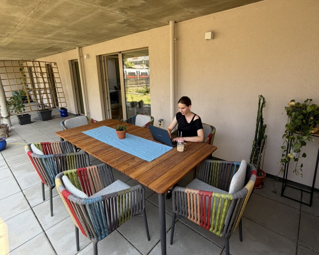 Eine Person am Laptop draußen auf der Terrasse. Ein großer Tisch mit bunten Stühlen und Pflanzen drumherum.